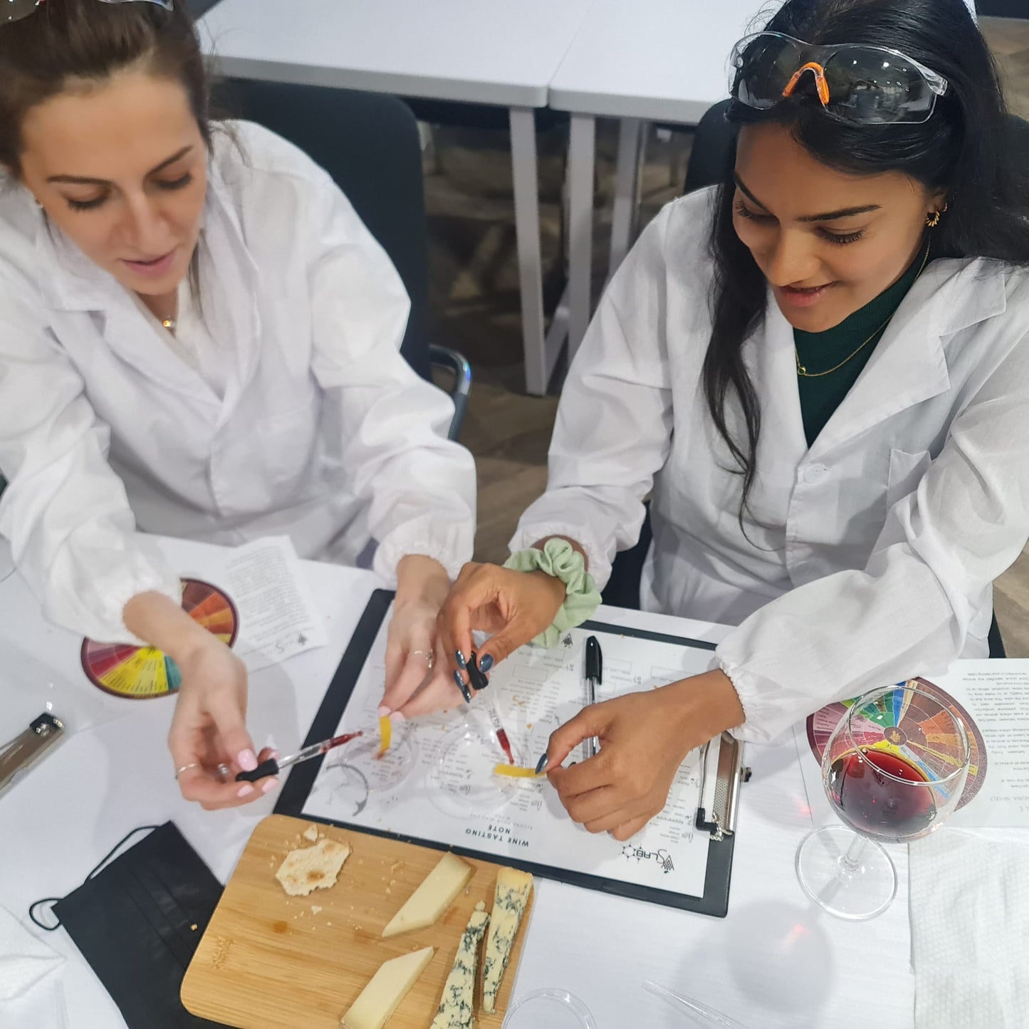 Two women in white lab coats conducting an experiment with wine and lab pipets, ph strips on a table.