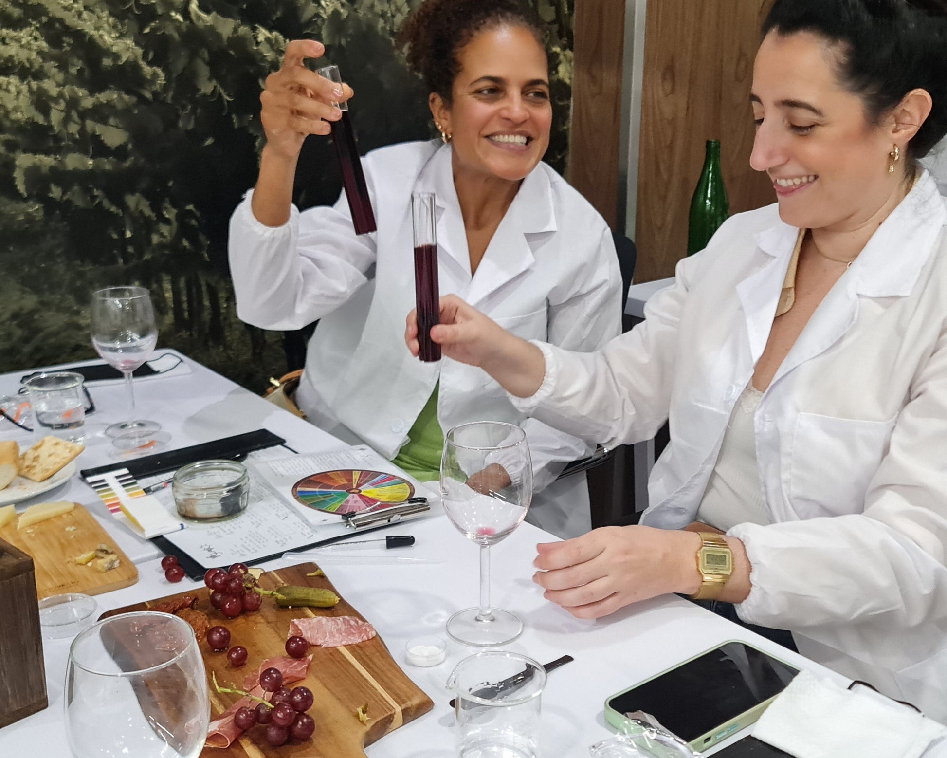 Two women at a dining table with wine glasses, cheese and charcuterie boards and lab testing tubes of wine.