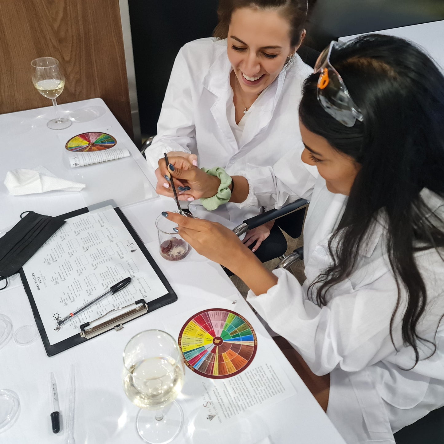 Two women in lab coats at a table with wine glasses, beakers, experimenting and color charts.