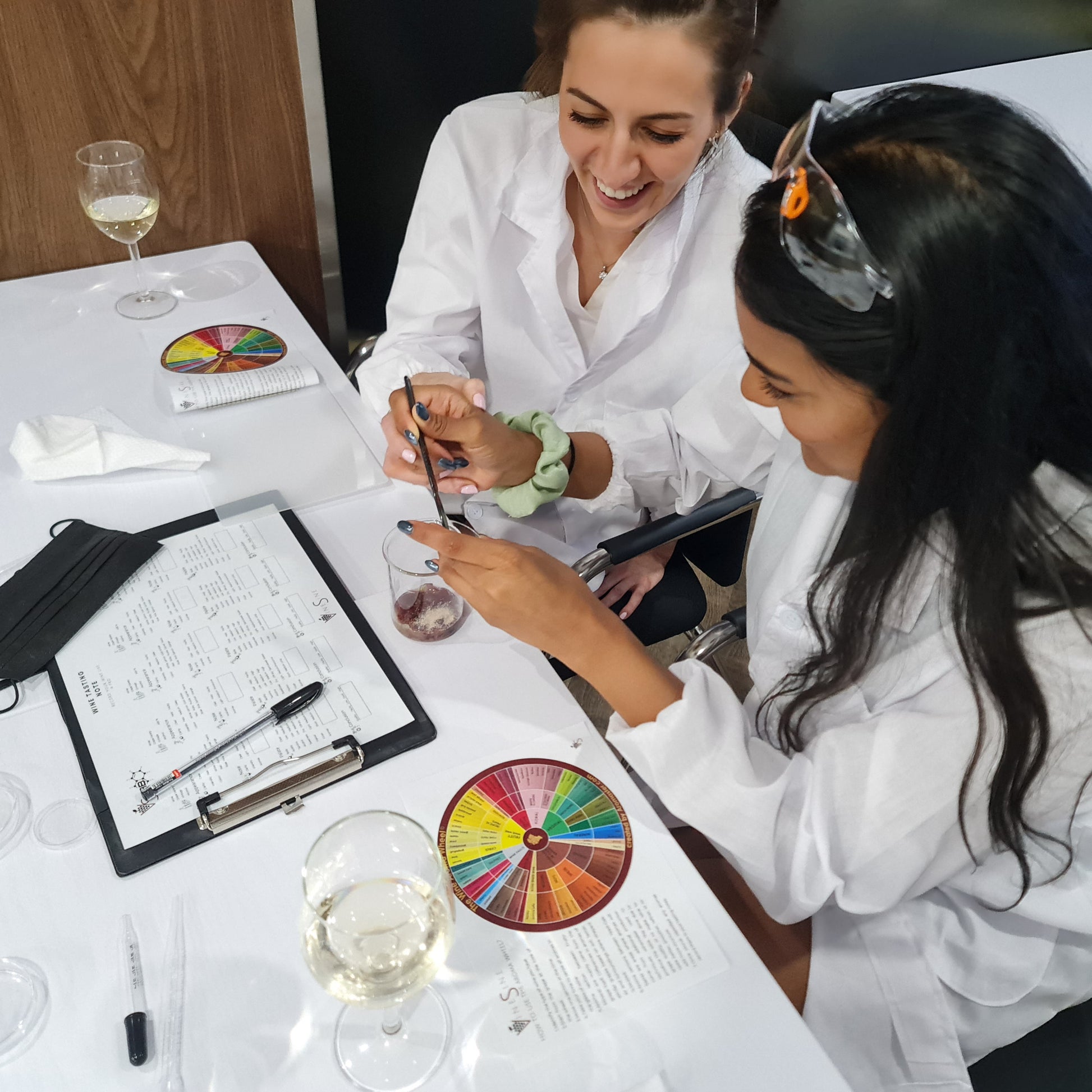 Two women in lab coats at a table with wine glasses, beakers, experimenting and color charts.